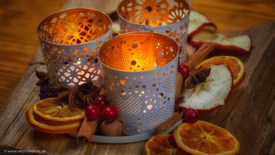 Drei Teelichthalter mit brennenden Kerzen warm erleuchtet inmitten von getrockneten Apfel- und Orangenscheiben, Sternanis, Zimtstanden, Beeren und Nüssen.