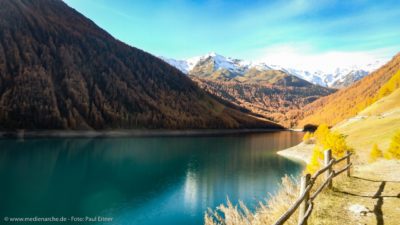 Ein azurblauer Bergsee im Herbst, gelbgefärbte Bäume am Ufer und schneebedeckte Berge im Hintergrund.