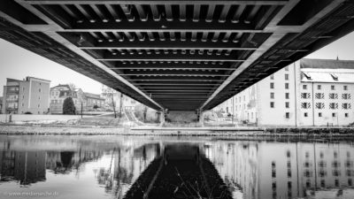 Unteransicht einer Brücke über einen ruhig dahinfließenden Fluss. Im Hintergrund historische Gebäude einer Stadt.