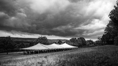 Ein großes Zelt, unter dem einige Menschen Unterschlupf vor einem aufziehenden Gewitter gefunden haben. Schwarz-weiß Fotografie.